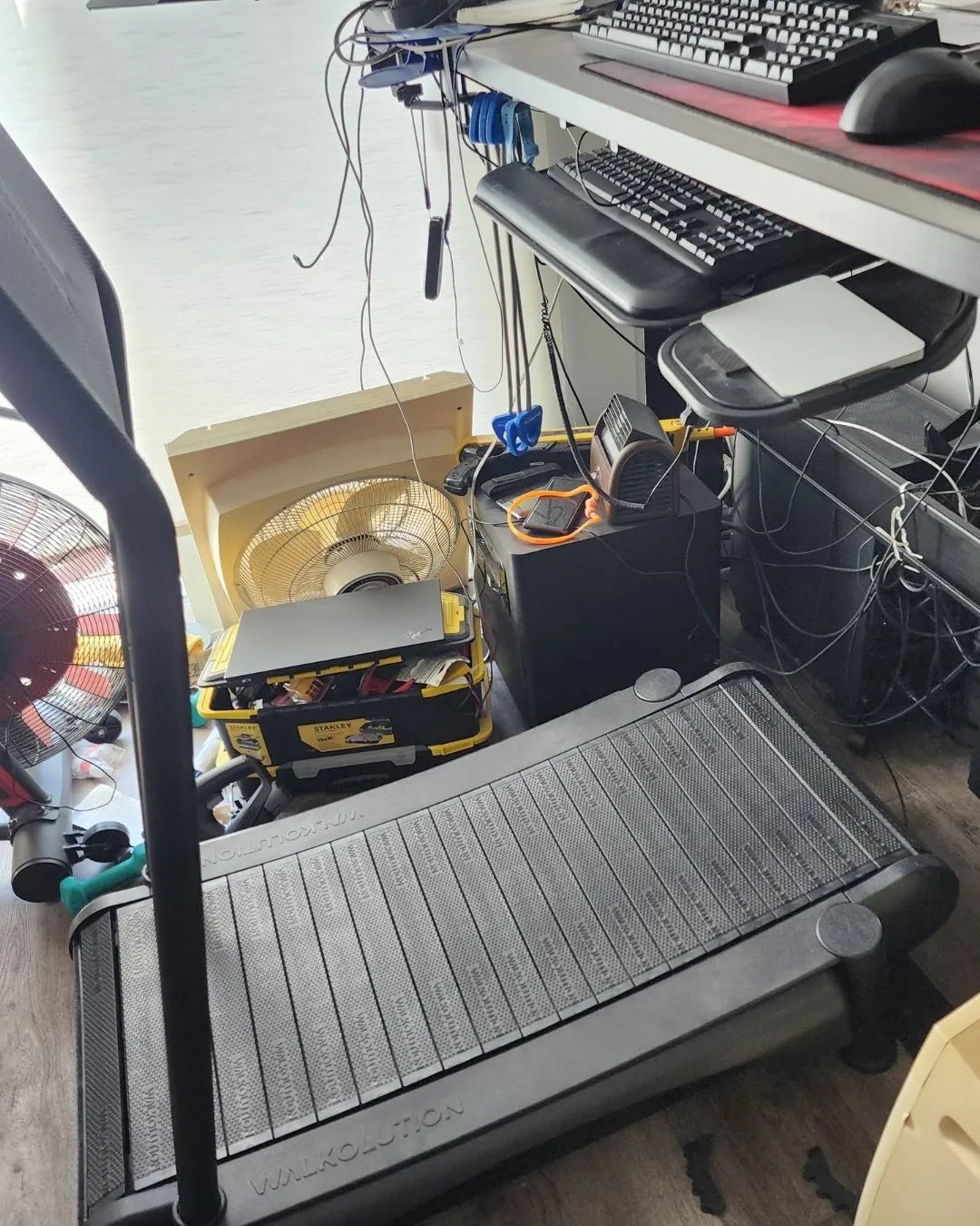 A treadmill is positioned in front of a cluttered desk with keyboards, wires, fans, and various electronics. A tool box and fan sit on the floor, and cables are tangled around the setup.