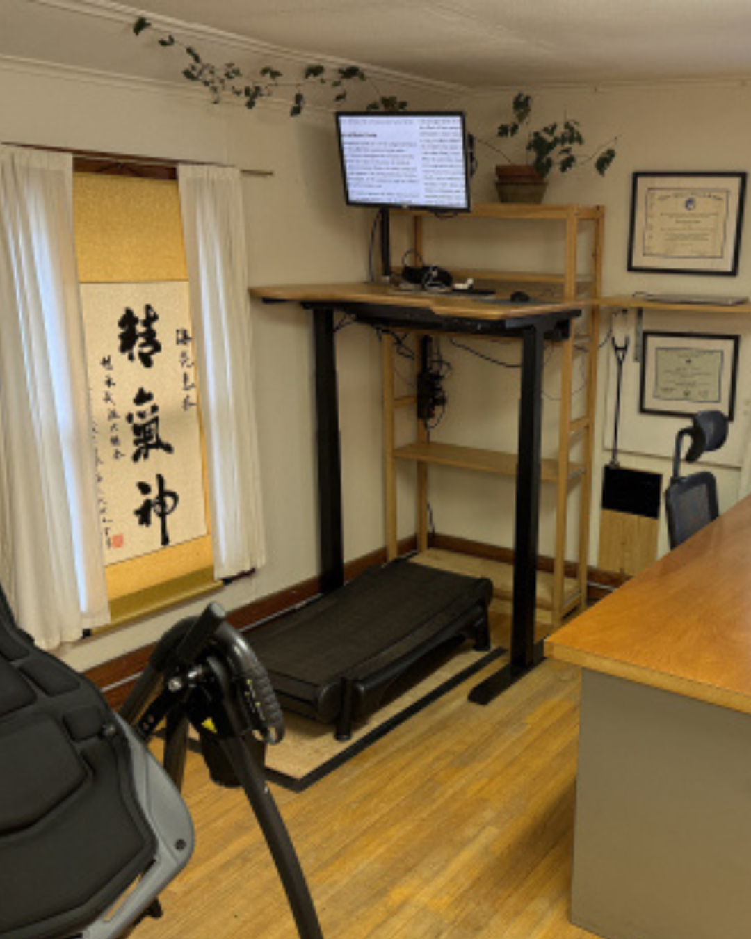 A home office features a treadmill desk with a monitor and keyboard on a wooden shelf. Diplomas hang on the wall, and a decorative scroll with Japanese characters is displayed beside a window with white curtains.